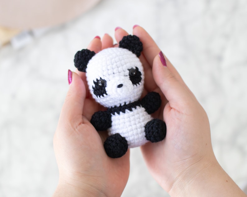 Op de afbeelding: Een klein gehaakt pandabeer speelgoed met zwart en wit bont. De panda heeft zwarte ogen en een zwarte neus.