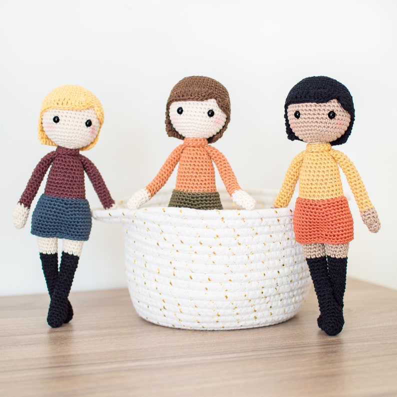 May include: Three crocheted dolls with different colored hair and outfits sitting in a white wicker basket. The dolls are wearing brown, orange, and yellow shirts and skirts.