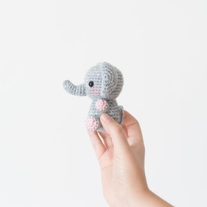May include: A small grey crocheted elephant with pink feet and a black eye. The elephant is being held in a person's hand.