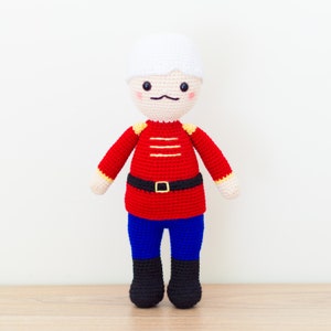 May include: A crocheted toy soldier with a white hat, red and yellow uniform, blue pants, and black boots.