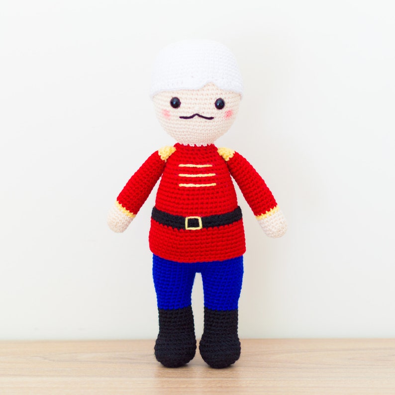 May include: A crocheted toy soldier with a white hat, red and yellow uniform, blue pants, and black boots.