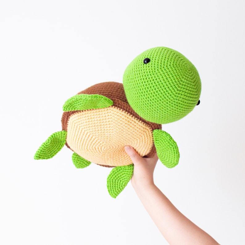 May include: A crocheted stuffed turtle toy with a green head and shell, brown legs, and a beige underside. The turtle is being held by a hand.