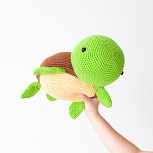 May include: A crocheted green and brown turtle plush toy with a yellow shell. The toy is being held by a hand.