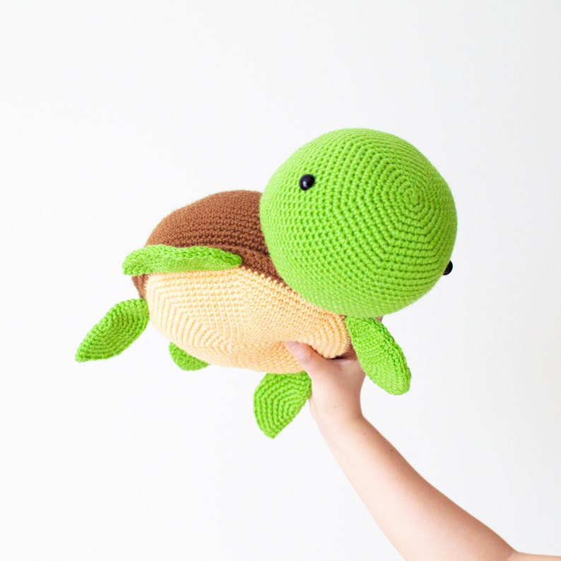 May include: A crocheted green and brown turtle plush toy with a yellow shell. The toy is being held by a hand.