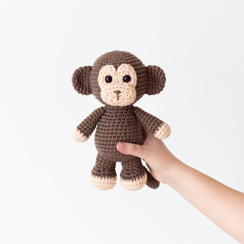 May include: A brown crocheted monkey toy with black eyes and a pink nose. The monkey is being held by a hand.