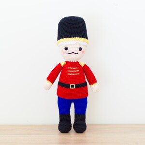 May include: A crocheted toy soldier with a red and yellow uniform, black hat, and blue pants. The toy has a friendly face with a mustache and rosy cheeks.
