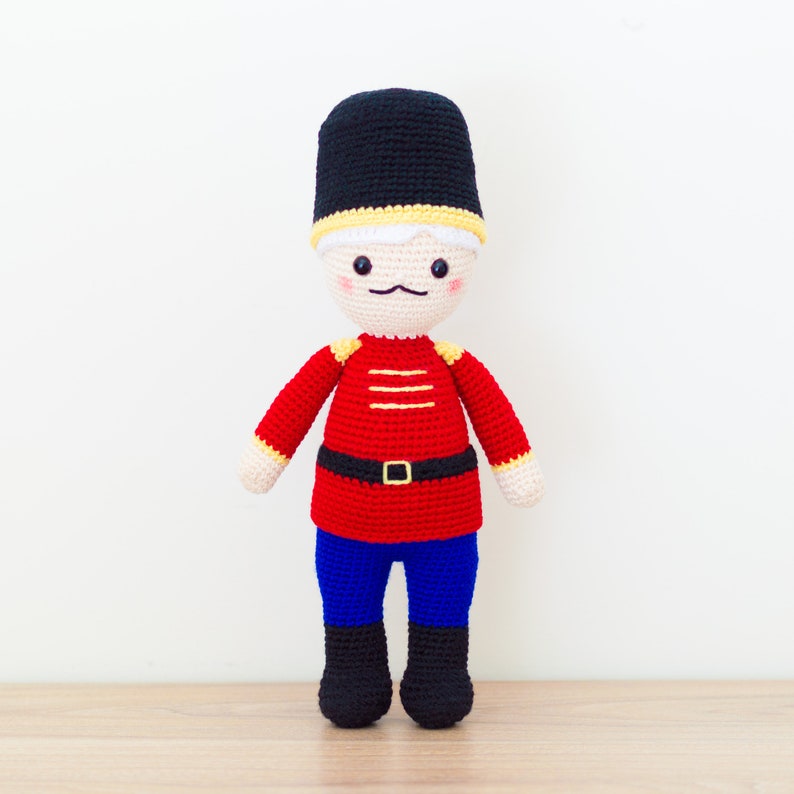 May include: A crocheted toy soldier with a red and yellow uniform, black hat, and blue pants. The toy has a friendly face with a mustache and rosy cheeks.