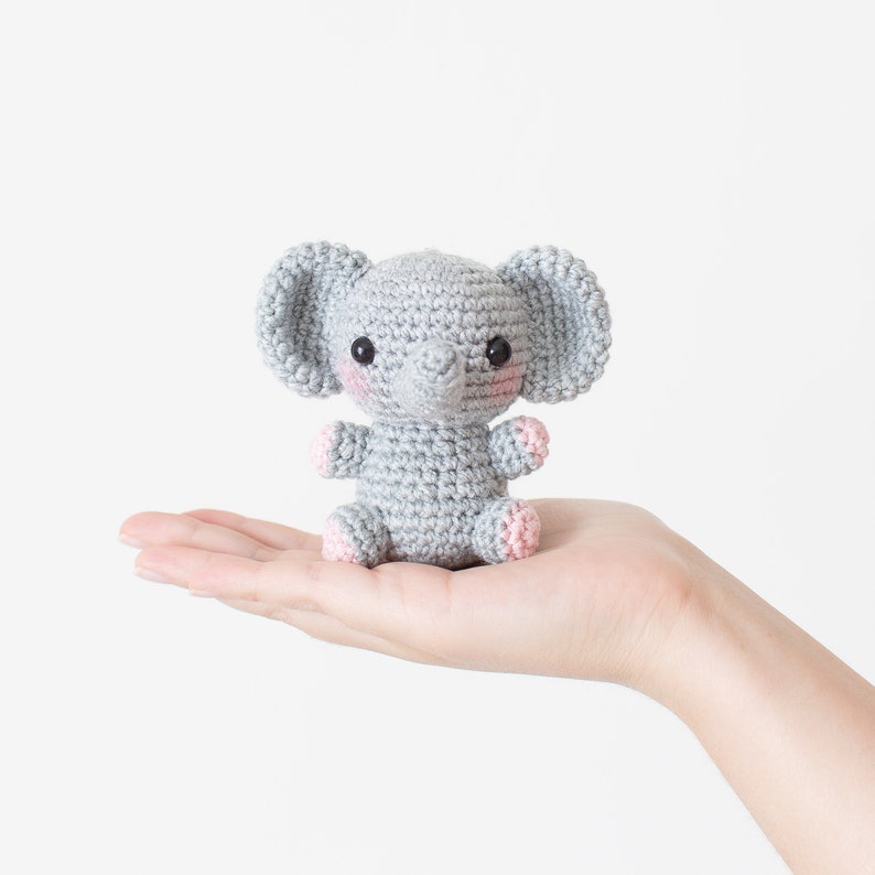 May include: A small grey crocheted elephant toy with pink cheeks and a grey trunk. The elephant is sitting in the palm of a hand.