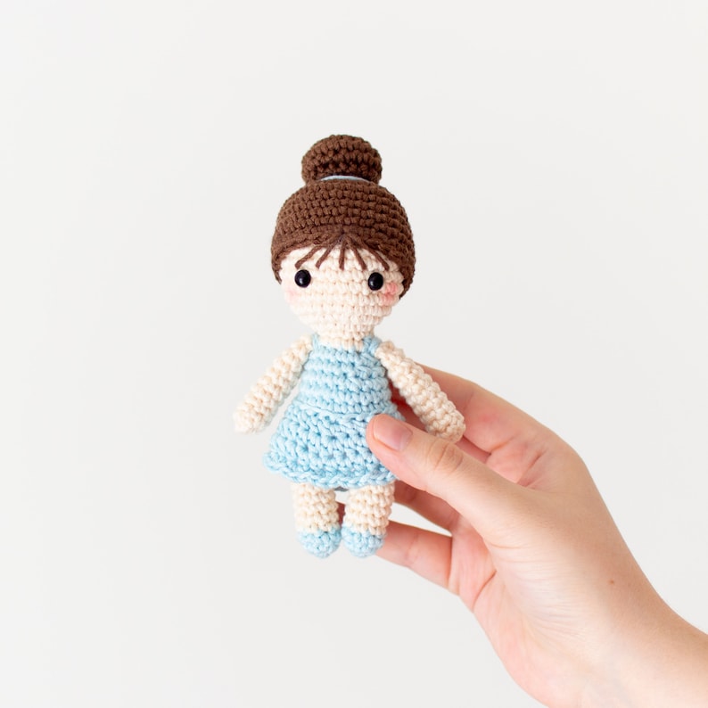 May include: A crocheted doll with brown hair in a bun, wearing a light blue dress. The doll has a sweet expression with black eyes and pink cheeks.