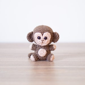May include: A brown crocheted monkey toy with black eyes and a light brown face. The monkey is sitting on a wooden surface.