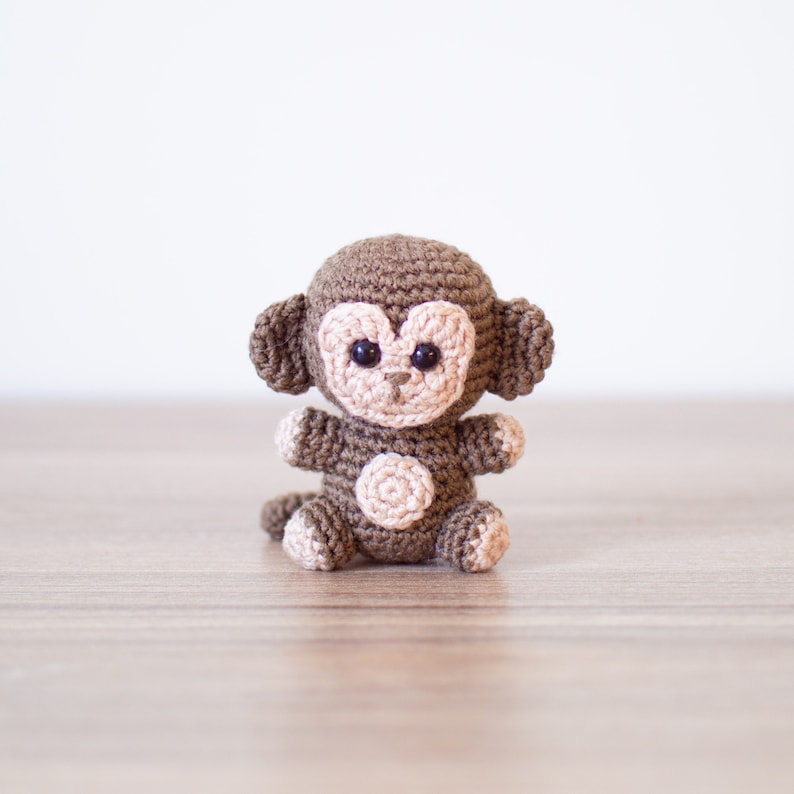 May include: A brown crocheted monkey toy with black eyes and a light brown face. The monkey is sitting on a wooden surface.