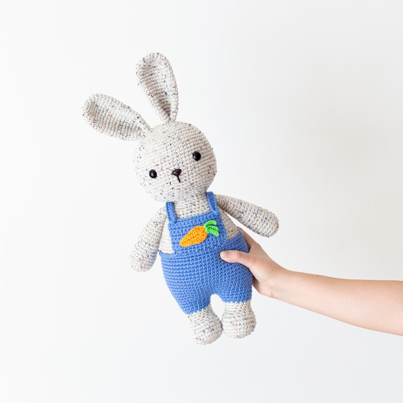 May include: A crocheted bunny toy wearing blue dungarees with a carrot patch. The bunny has white fur with speckles of colour and black eyes.