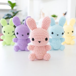May include: Five crocheted bunny plushes in different pastel colors: green, purple, pink, blue, and yellow. The bunnies have black eyes and are sitting on a white surface.