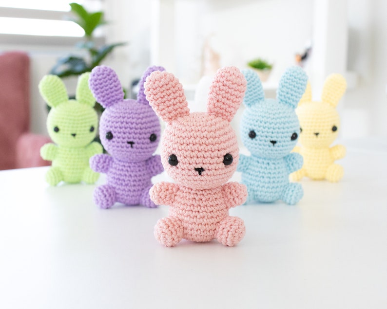 May include: Five crocheted bunny plushes in different pastel colors: green, purple, pink, blue, and yellow. The bunnies have black eyes and are sitting on a white surface.
