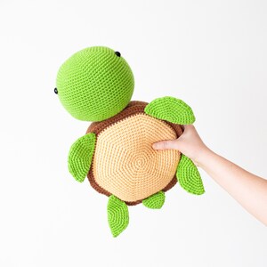 May include: A crocheted green and brown turtle toy with black eyes. The turtle is being held by a hand.