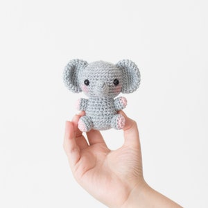 May include: A small grey crocheted elephant toy with pink accents on the ears and feet. The elephant has black eyes and a small grey trunk.