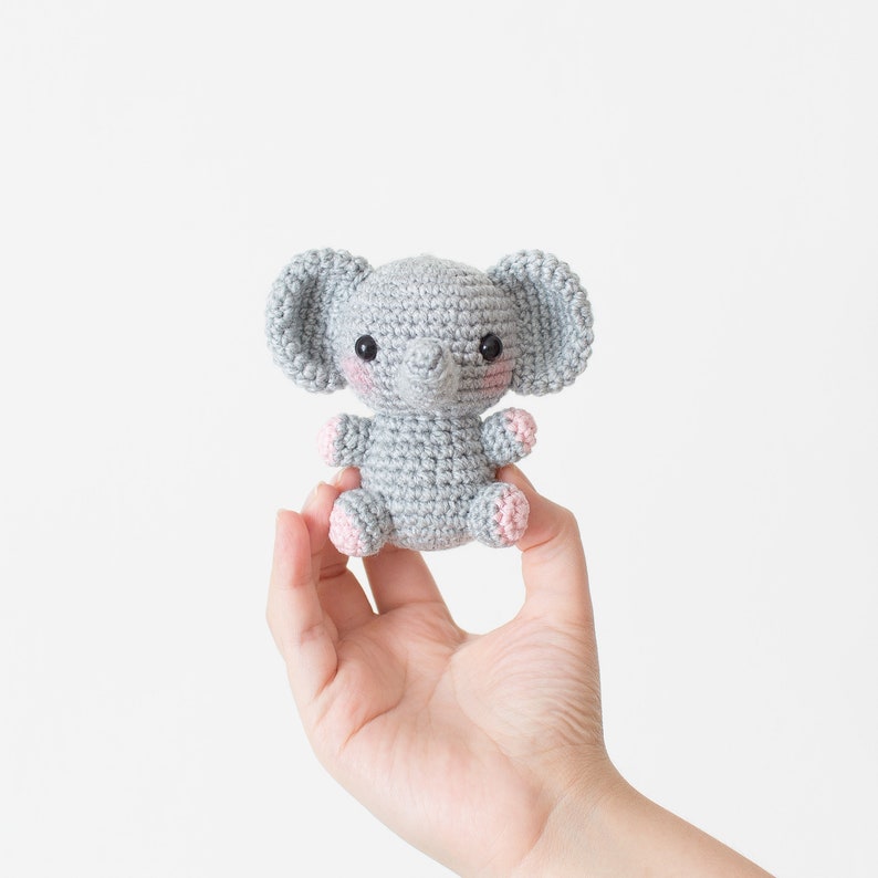 May include: A small grey crocheted elephant toy with pink accents on the ears and feet. The elephant has black eyes and a small grey trunk.