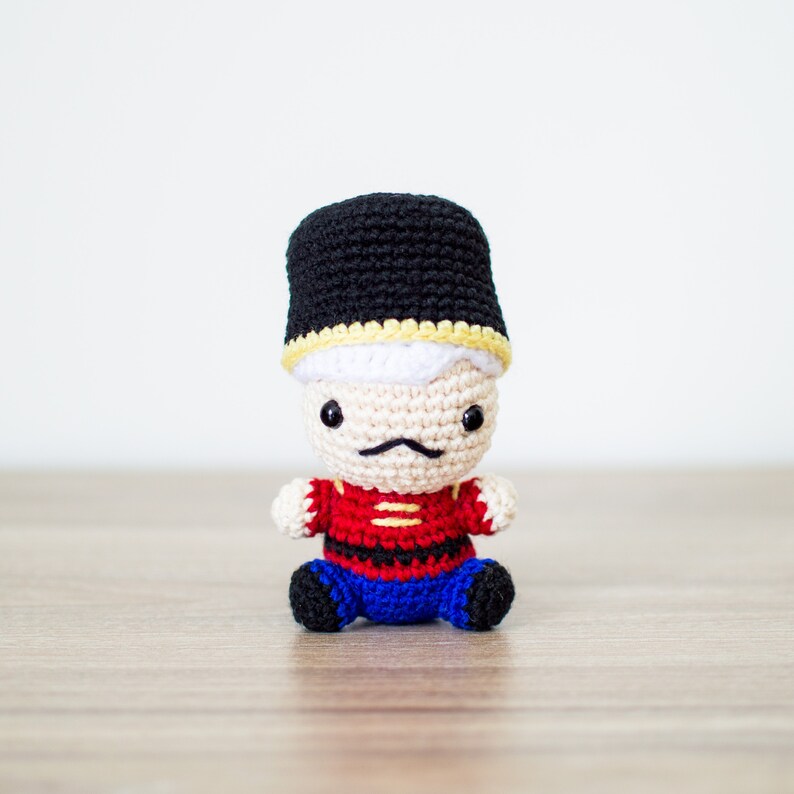 May include: A handmade crochet doll of a toy soldier. The doll has a black hat with yellow trim, a red jacket with gold accents, blue pants, and black boots. The doll has a white face, a black mustache, and black eyes.