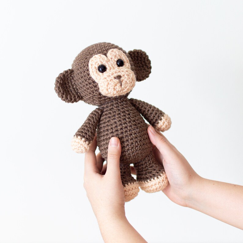 May include: A brown crocheted monkey toy with black eyes and a light brown nose. The monkey is being held by two hands.