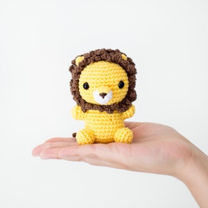 May include: A small crocheted lion toy with a brown mane and yellow body. The lion has black eyes and a white nose.
