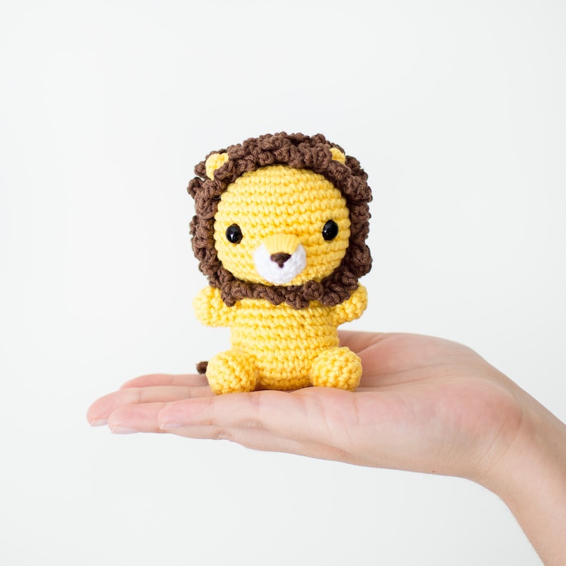 May include: A small crocheted lion toy with a brown mane and yellow body. The lion has black eyes and a white nose.