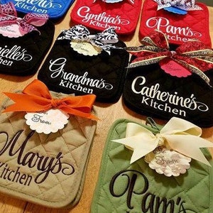 May include: A collection of ten personalised kitchen pot holders with various names embroidered on them. The pot holders are in different colours, including red, blue, black, green, and beige. Each pot holder has a bow and a tag attached to it.