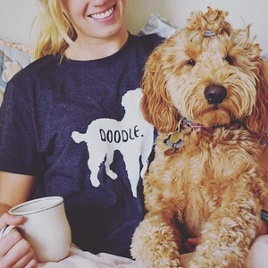 May include: A dark blue t-shirt with a white doodle dog graphic and the word "DOODLE." A light brown Goldendoodle dog is in the image. The dog has a collar with tags. A white mug with a blue rim is also in the image.