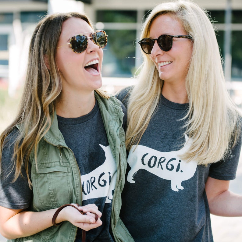 May include: Two women wearing gray t-shirts with a white corgi dog graphic and the word "CORGI." One woman wears a green vest and sunglasses. The other woman wears sunglasses. They are smiling and laughing.