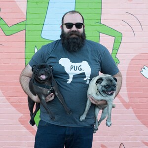 May include: A man wearing a gray t-shirt with a white pug graphic and the word "Pug." He is holding a black pug and a tan pug. The background features a colorful mural with a cartoon character.