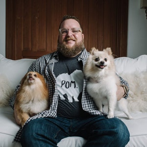 May include: A person wearing glasses and a gray t-shirt with the word "POM." and a bear graphic, holding two Pomeranian dogs. The person is sitting on a white couch, wearing a plaid shirt and blue jeans.