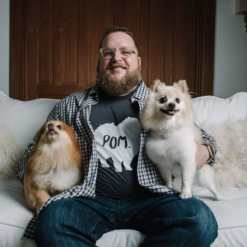 May include: A person wearing glasses and a gray t-shirt with the word "POM." and a bear graphic, holding two Pomeranian dogs. The person is sitting on a white couch, wearing a plaid shirt and blue jeans.