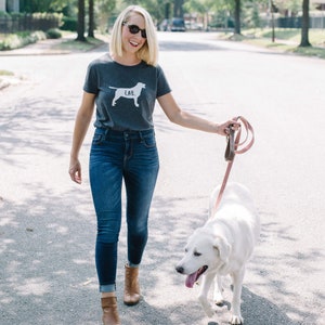 May include: A dark gray t-shirt with a white Labrador Retriever graphic and the word "LAB." A person is walking a white dog on a leash. The person is wearing blue jeans and brown boots. The leash is red and white striped.