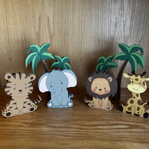 Peut inclure: Quatre figurines d'animaux en bois avec des visages de dessin anim&eacute;, dont un tigre, un &eacute;l&eacute;phant, un lion et une girafe. Les figurines sont assises sur une surface en bois devant des palmiers.