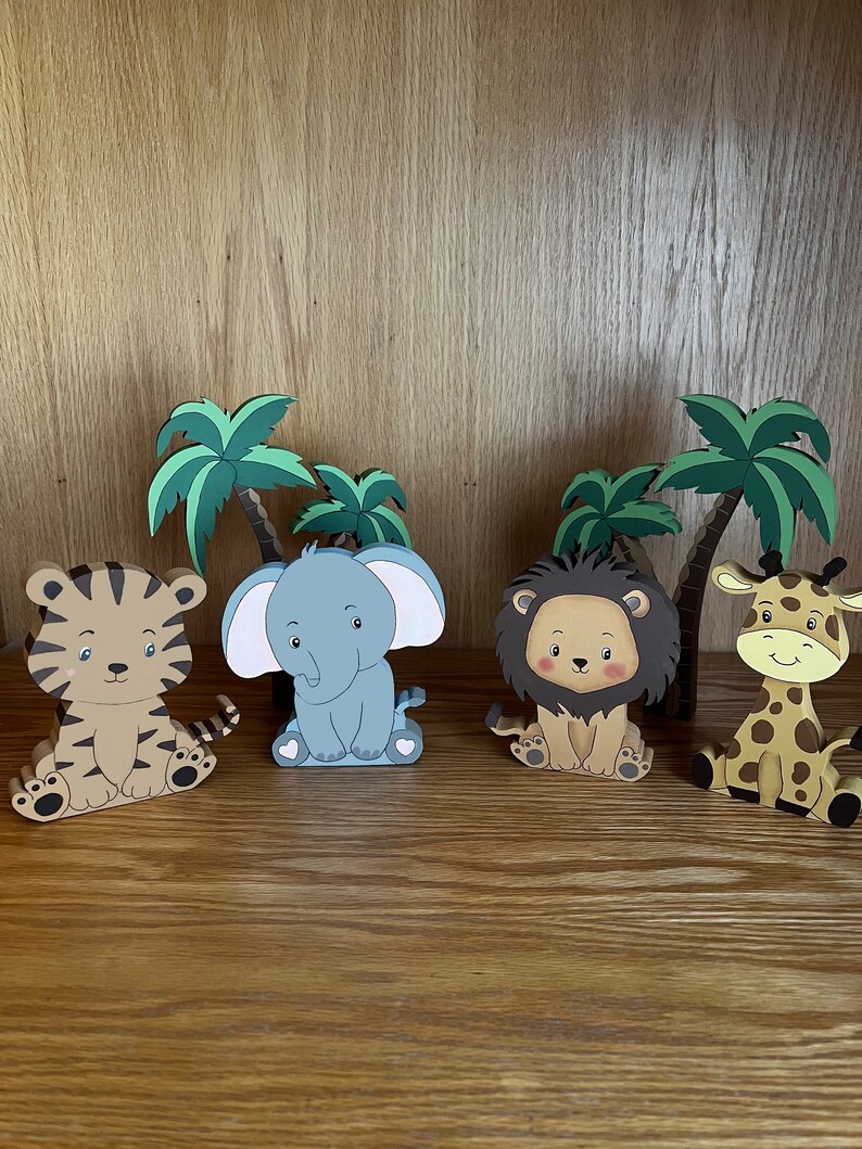 Peut inclure: Quatre figurines d'animaux en bois avec des visages de dessin anim&eacute;, dont un tigre, un &eacute;l&eacute;phant, un lion et une girafe. Les figurines sont assises sur une surface en bois devant des palmiers.