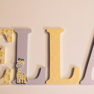 May include: Wooden letters spelling the name "ELLA" in yellow and grey with a giraffe pattern on the "E" and "L" and a small giraffe figurine on the "L".