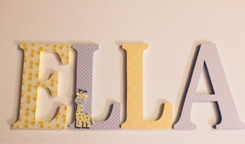 May include: Wooden letters spelling the name "ELLA" in yellow and grey with a giraffe pattern on the "E" and "L" and a small giraffe figurine on the "L".