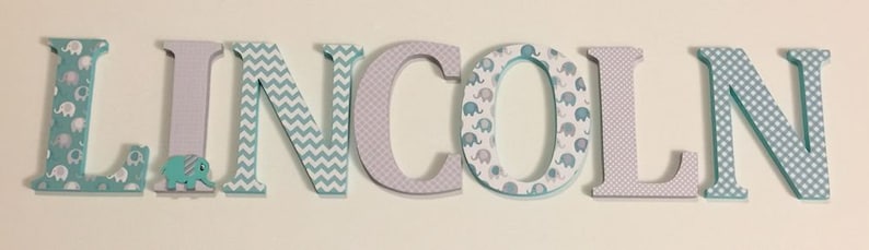 May include: Wooden letters spelling the name "LINCOLN" in light blue, gray, and white. The letters are decorated with a variety of patterns, including elephants, chevron, and polka dots.