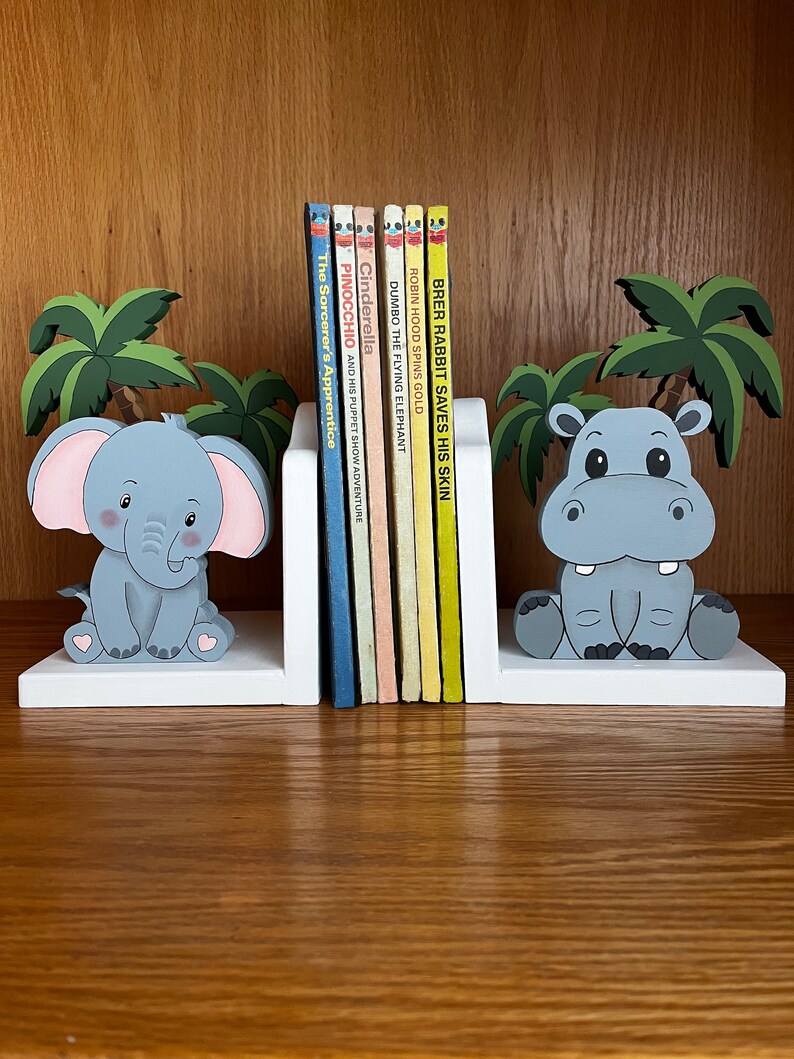 Peut inclure: Des serre-livres en bois blanc avec un &eacute;l&eacute;phant gris et un hippopotame gris. Les serre-livres sont d&eacute;cor&eacute;s de palmiers. Les serre-livres tiennent une rang&eacute;e de livres pour enfants color&eacute;s avec des titres comme "Pinocchio", "Cendrillon" et "Dumbo l'&eacute;l&eacute;phant volant".
