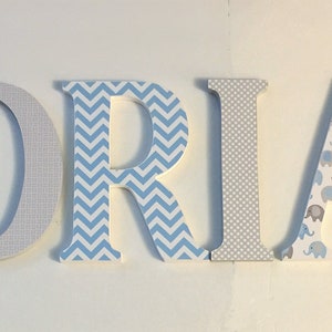 May include: Wooden letters spelling the name "ADRIAN" in blue, gray, and white. The letters are decorated with different patterns, including elephants, chevron, and geometric designs.