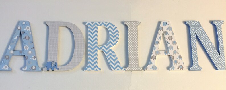 May include: Wooden letters spelling the name "ADRIAN" in blue, gray, and white. The letters are decorated with different patterns, including elephants, chevron, and geometric designs.