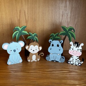 Peut inclure: Quatre figurines d'animaux en bois avec des palmiers derri&egrave;re elles. Les animaux sont un koala bleu, un singe brun, un hippopotame bleu et un z&egrave;bre noir et blanc. Tous les animaux sont assis.