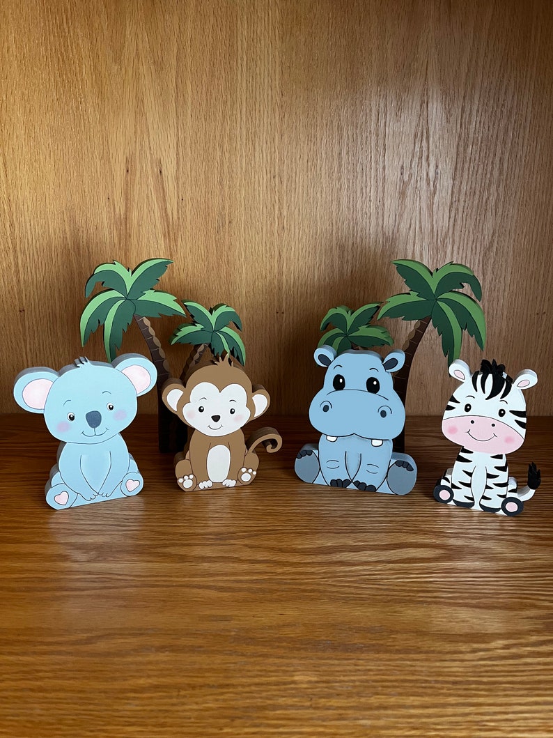 Peut inclure: Quatre figurines d'animaux en bois avec des palmiers derri&egrave;re elles. Les animaux sont un koala bleu, un singe brun, un hippopotame bleu et un z&egrave;bre noir et blanc. Tous les animaux sont assis.