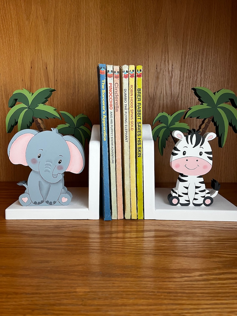 Peut inclure: Des serre-livres en bois blanc avec un &eacute;l&eacute;phant et un z&egrave;bre peints. L'&eacute;l&eacute;phant est gris avec des oreilles roses et est assis sur une base blanche. Le z&egrave;bre est noir et blanc avec un nez rose et est assis sur une base blanche. Les serre-livres tiennent une rang&eacute;e de livres pour enfants color&eacute;s avec des titres comme "Pinocchio", "Cendrillon" et "Dumbo l'&eacute;l&eacute;phant volant".