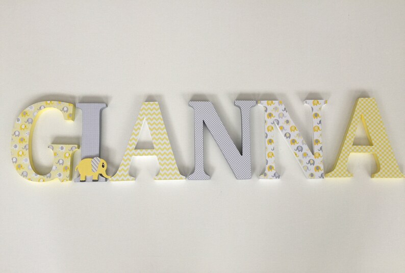 May include: Wooden letters spelling the name "GIANNA" in yellow and gray. The letters are decorated with different patterns, including elephants, chevron, and polka dots.