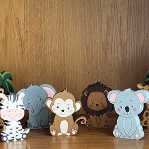 Peut inclure: Un ensemble de sept figurines d'animaux en bois, comprenant un tigre, un z&egrave;bre, un &eacute;l&eacute;phant, un singe, un lion, un koala et une girafe. Les figurines sont peintes dans des couleurs vives et ont un style cartoon. Elles sont toutes assises sur une surface en bois.