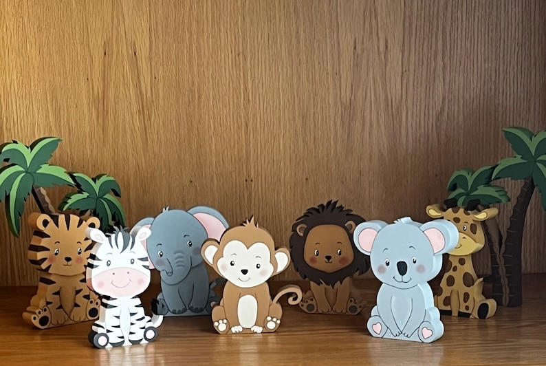 Peut inclure: Un ensemble de sept figurines d'animaux en bois, comprenant un tigre, un z&egrave;bre, un &eacute;l&eacute;phant, un singe, un lion, un koala et une girafe. Les figurines sont peintes dans des couleurs vives et ont un style cartoon. Elles sont toutes assises sur une surface en bois.