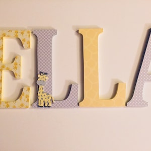 May include: Wooden letters spelling the name "ELLA" in yellow and grey with a giraffe cutout on the "L".