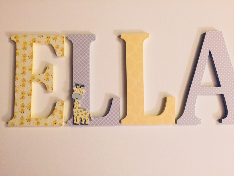 May include: Wooden letters spelling the name "ELLA" in yellow and grey with a giraffe cutout on the "L".