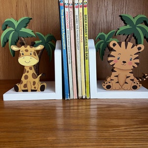 Peut inclure: Une paire de serre-livres en bois blanc avec une figurine de girafe et une figurine de tigre. Les serre-livres soutiennent une rang&eacute;e de livres pour enfants avec des titres comme "Cendrillon", "Pinocchio" et "Dumbo l'&eacute;l&eacute;phant volant".