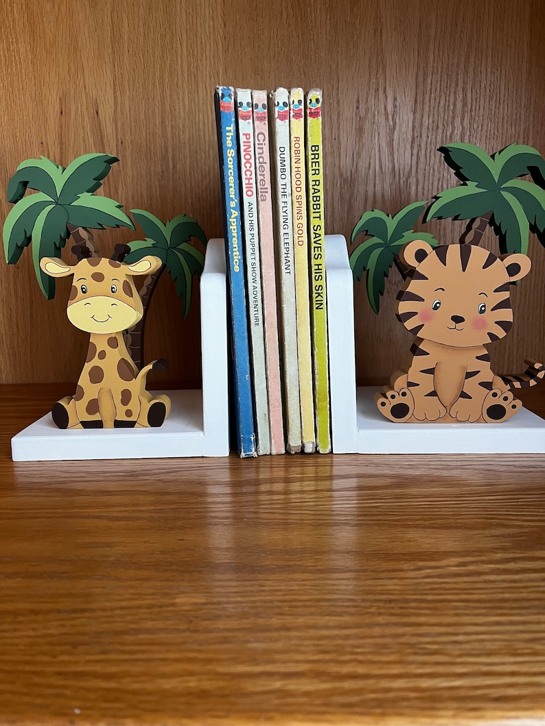 Peut inclure: Une paire de serre-livres en bois blanc avec une figurine de girafe et une figurine de tigre. Les serre-livres soutiennent une rang&eacute;e de livres pour enfants avec des titres comme "Cendrillon", "Pinocchio" et "Dumbo l'&eacute;l&eacute;phant volant".